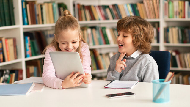Two primary school kids sitting together at desk in classroom or library interior, using digital tablet computer
