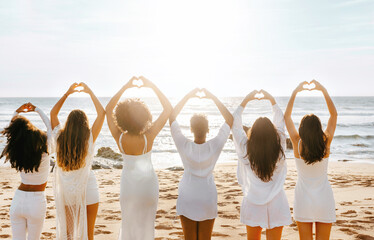 Back view of young women doing hearth symbol with hands, standing in line by ocean shore at sunset,...