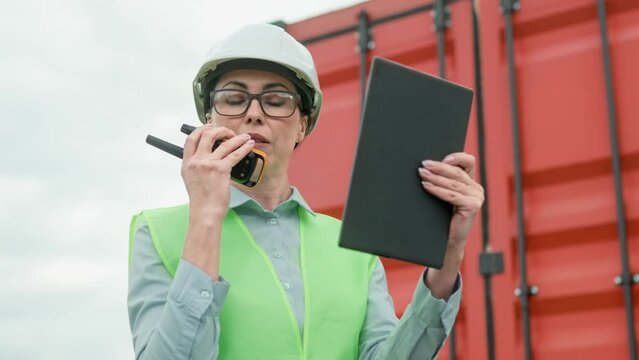 Busy Caucasian woman worker communicating with partners on walkie-talkie and using tablet device. Beautiful female with safety helmet and vest standing near red container. Transportation company.