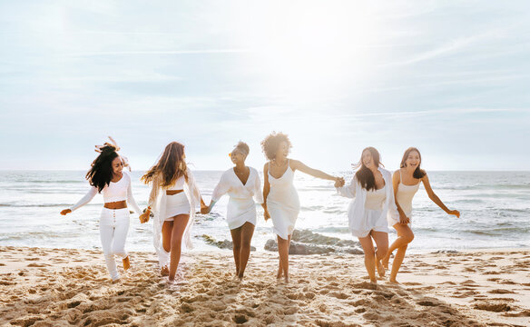 Group of joyful diverse women enjoying hen party, running and having fun on the beach at coastline, full length