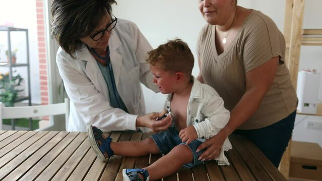female senior pediatrician doctor, listening to crying little boy next to his mom