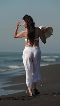 Rear view of woman walking along sandy beach on Pacific Coast during summer beach holidays. 50-year-old Caucasian ethnicity sensual female in bikini top, cotton sheer capris and straw sun hat in hand