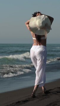 Rear view of sexy unrecognizable barefoot woman walking on beach with her hands raised behind head and holding straw sun hat. Full length sensuality barefoot female in white see-through capri pants
