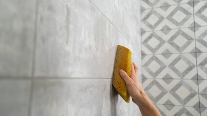 Close-up of wiping after the process of grouting cracks on a tile wall. A man's hand wipes a tiled wall with a sponge after repair work. A man cleans the bathroom with a sponge. - obrazy, fototapety, plakaty