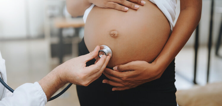 Pregnant african woman has appointment with doctor at clinic. Male gynaecologist OB GYN medic specialist with stethoscope listens to baby's heartbeat in mother's belly. Pregnancy, health care