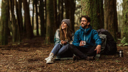 Smiling young caucasian couple in jackets sits in forest, drinking cups of tea, enjoy travel, vacation and hot coffee