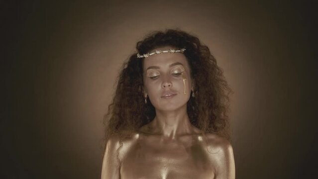 Attractive curly haired young woman with golden makeup tears and looking at the camera shyly, golden paint and accessories, crown of thornes on the head, close up video.