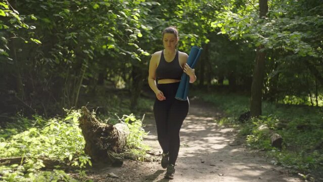 A beautiful girl walks with a yoga mat through the forest to do fitness. A healthy lifestyle of playing sports and practicing yoga asanas. High quality 4k footage