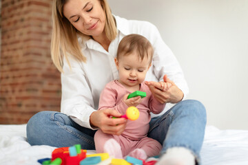 Mother sitting with baby daughter on bed and playing toys together, educating games for infants and toddlers