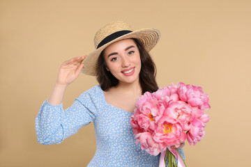 Beautiful young woman in straw hat with bouquet of pink peonies against beige background