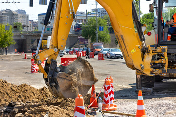 The bucket of a construction excavator digs a trench on the road against the backdrop of a city street. - obrazy, fototapety, plakaty