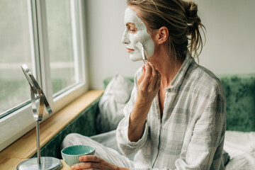 A woman applies a clay mask to her face with a brush while looking in the mirror.