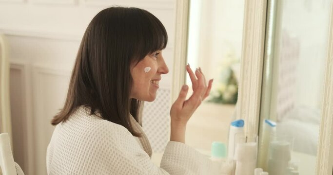 Content woman is seen using facial lotion near the vanity. Soft lighting accentuates the tranquil ambiance, creating a comfortable and soothing space for her skincare routine.