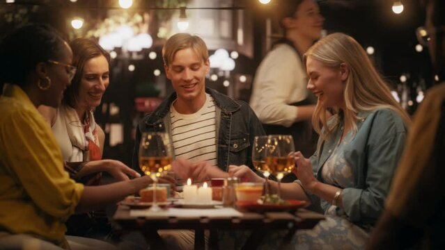 Young Multiethnic Friends Enjoying a Night Out in a Cafe. Waiter Bringing a Delicious Italian Pizza. Young Men and Women Having Fun Conversations on a Terrace, Enjoying Tasty Food and Beverages