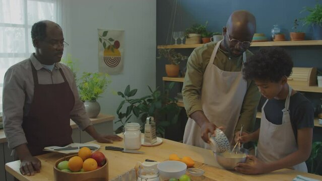 Medium long shot of black father adding liquid ingredient to bowl with cake batter mixed by his little son while granddad reading recipe from family cookbook in kitchen