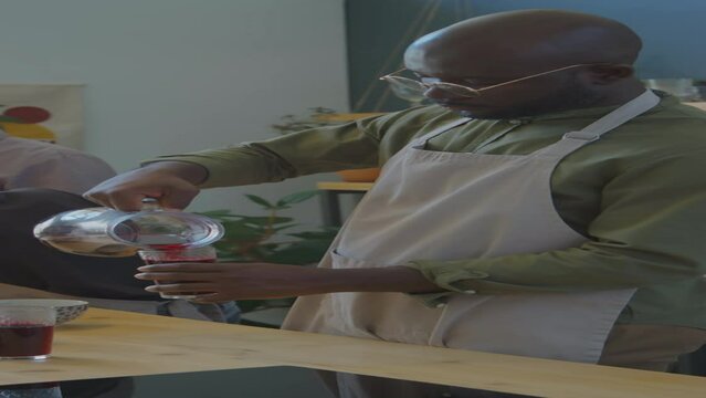 Vertical shot of African American man pouring Zobo tea into glass while preparing traditional food with family members in kitchen at home