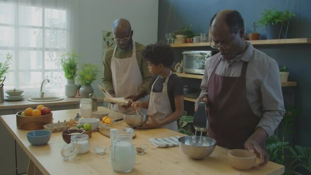 Senior black man using mixer to make cake batter while his son and grandson using cookbook and whisking ingredients beside at kitchen table