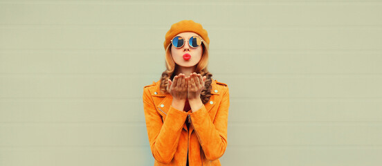 Autumn portrait of stylish beautiful young woman model blowing her lips sends sweet kiss wearing orange french beret hat, jacket and round sunglasses on gray background
