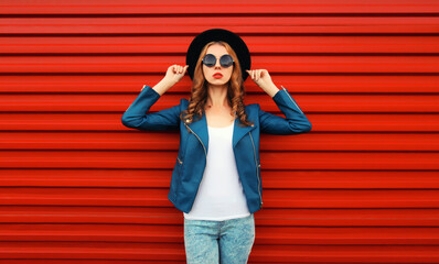 Portrait of beautiful young woman wearing blue leather jacket, sunglasses, round hat on red wall background