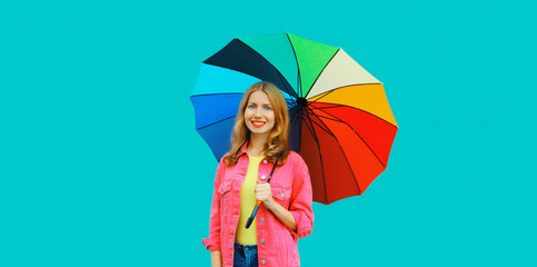 Autumn portrait of stylish happy smiling young woman holding colorful umbrella wearing pink jacket on blue background
