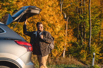 a smiling man with a backpack on his back closes the trunk of the car