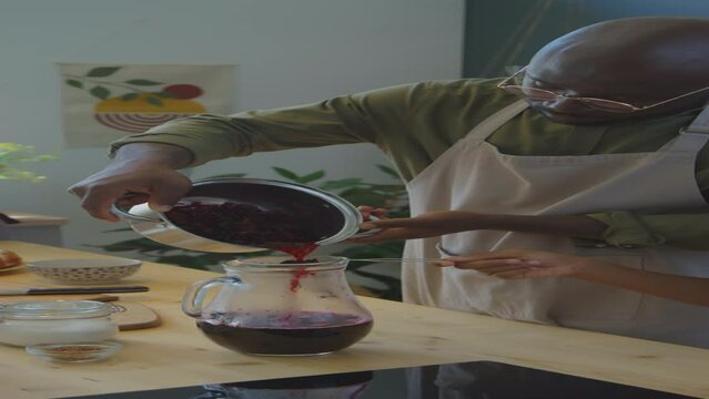 Vertical shot of African American man straining Zobo tea into glass pitcher with help of little son while cooking together in kitchen at home