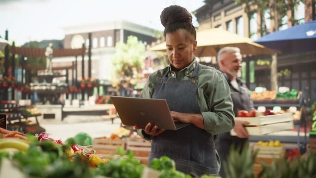 African Farmer Using Laptop Computer on a City Square with Vendors Selling Fresh Organic Agricultural Products. Middle Aged Black Female Managing Business Operations and Communications with Suppliers