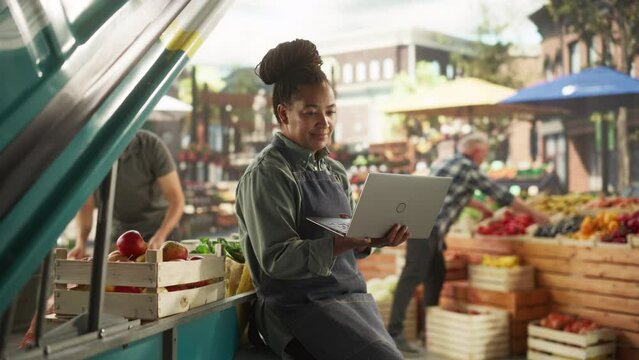 Multiethnic African Businesswoman Working on Laptop Computer, Preparing for an International Food Festival on a City Square. Female Sitting Next to a Street Vendor Booth with Organic Farm Products