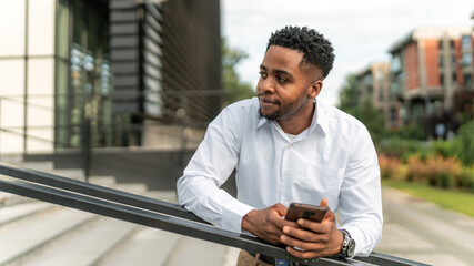 A portrait of a handsome Black businessman leaning against a railing during his break, holding a phone in his hand. 