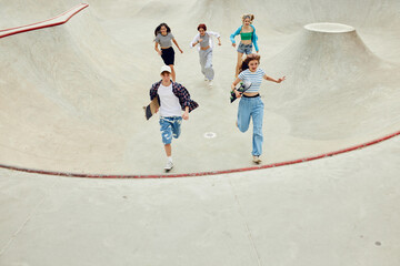 Group of teens in casual clothes, boy and girls running with skate on skateboard ramp. Activity and fun. Concept of youth culture, sport, dynamic, extreme, hobby, action and motions, friendship