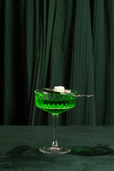 Green cocktail in a crystal glass with a sugar cube on a silver spoon on a dark green background
