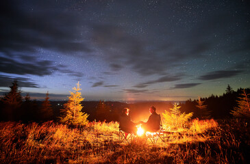 Happy couple staring at starry sky. Young people sitting in chairs and warming by campfire....