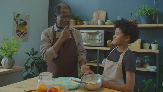 African American grandfather putting batter on nose of cute little grandson while baking together cake in kitchen at home and having fun