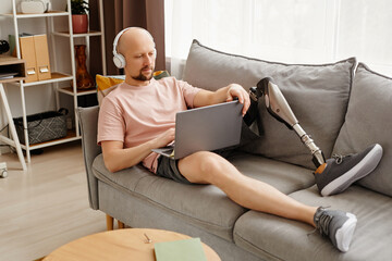 Full length portrait of adult man with prosthetic leg using laptop while lying on sofa at home and wearing headphones