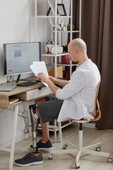 Full length side view of man with prosthetic leg working from home and reading documents at desk