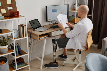 Portrait of bald adult man with prosthetic leg working from home and reading documents at desk