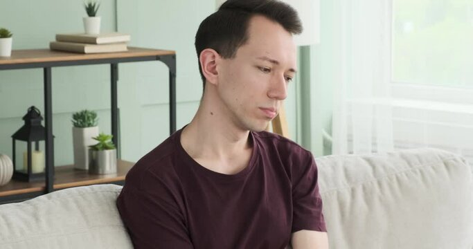 In a poignant scene, an upset Caucasian man is sitting on the sofa in the living room. His troubled expression and slouched posture reflect his distress and sadness.