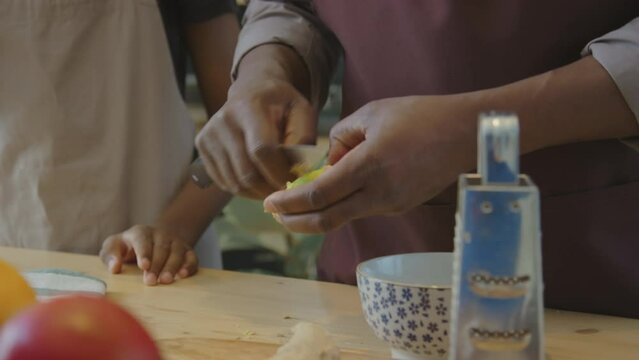 Cropped shot of African American man in apron peeling fresh ginger root with knife while cooking with kid in kitchen at home