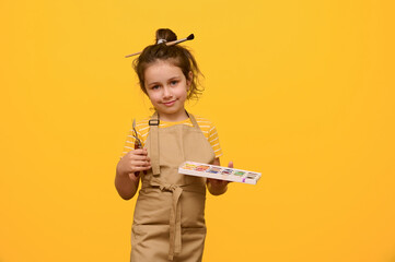 Studio portrait beautiful little girl, painter artist smiles looking at camera, holds painting tools and a palette with watercolors, isolated over yellow studio background. Kids. Creativity. Art class