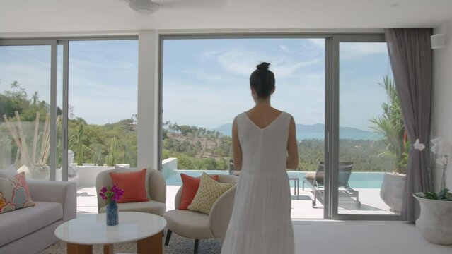 Stylish woman in white summer dress put off her beach bag and walking out to lounge area open terrace and swimming pool on jungle background, tropical holiday concept.