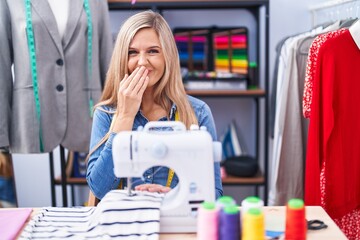 Blonde woman dressmaker designer using sew machine laughing and embarrassed giggle covering mouth...