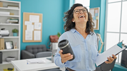 Middle age hispanic woman business worker holding coffee and clipboard dancing at the office
