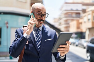 Young hispanic man executive talking on smartphone using touchpad at street