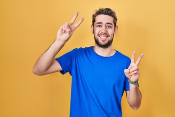 Hispanic man with beard standing over yellow background smiling with tongue out showing fingers of...