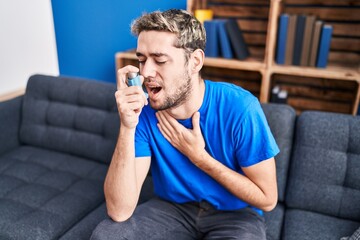 Young man using inhaler sitting on sofa at home - obrazy, fototapety, plakaty