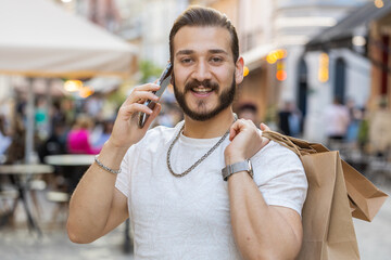 Joyful smiling bearded young man shopaholic consumer after shopping sale with full bags with gifts outdoors. Guy tourist traveler walking along the urban sunshine city street road. Town lifestyles