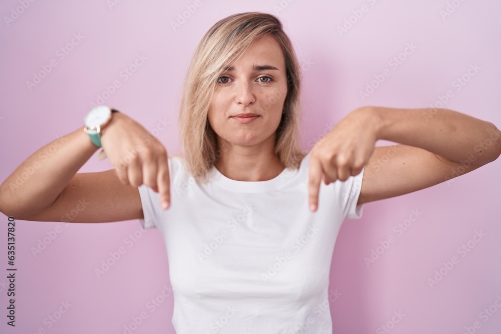 Sticker Young blonde woman standing over pink background pointing down looking sad and upset, indicating direction with fingers, unhappy and depressed.