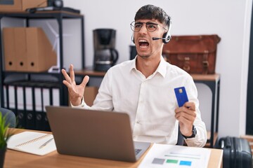 Young hispanic man working using computer laptop holding credit card crazy and mad shouting and yelling with aggressive expression and arms raised. frustration concept.