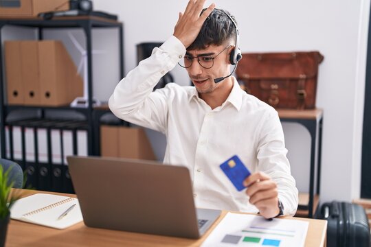 Young hispanic man working using computer laptop holding credit card surprised with hand on head for mistake, remember error. forgot, bad memory concept.