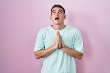 Handsome hispanic man standing over pink background begging and praying with hands together with...